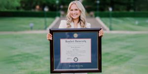 Smiling woman on campus holding a large Samford University diploma frame.