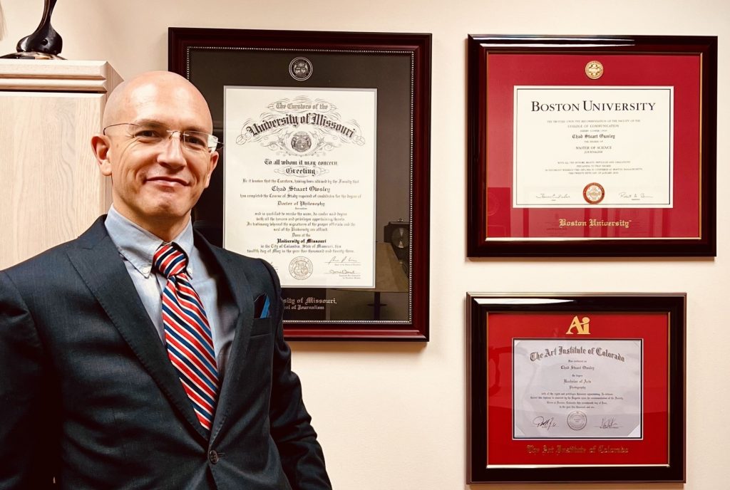 Man in suit standing next to wall with University of Missouri, Art Institute of Colorado and Boston University diploma frames.