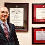 Man in suit standing next to wall with University of Missouri, Art Institute of Colorado and Boston University diploma frames.