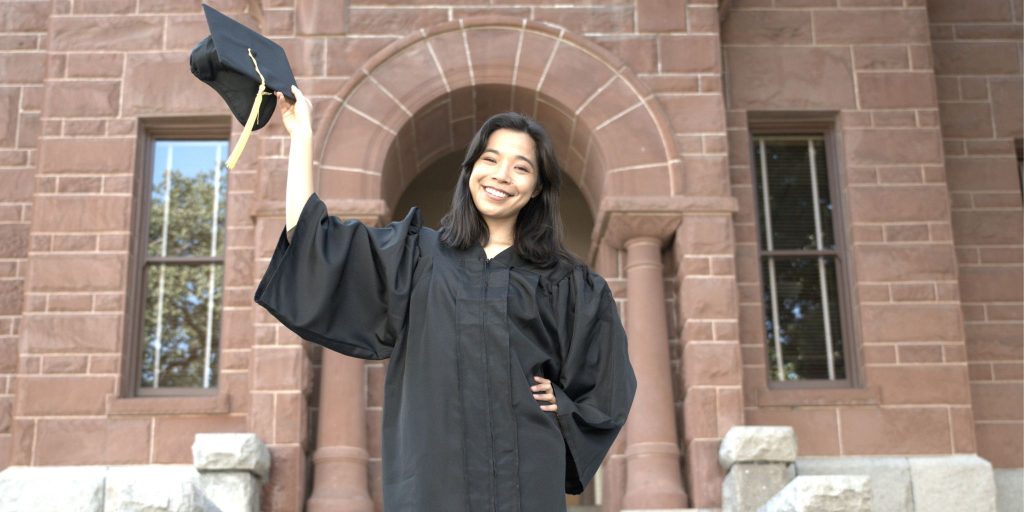 Smiling graduate in front of brick campus building holding up her graduation cap.