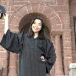 Smiling graduate in front of brick campus building holding up her graduation cap.