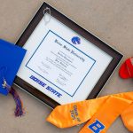 Boise State diploma frame on pavement next to graduation cap, red shoes, and a yellow graduation stole.