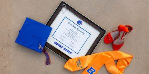 Boise State diploma frame on pavement next to graduation cap, red shoes, and a yellow graduation stole.