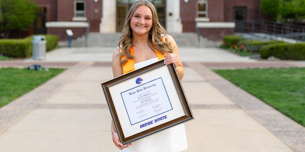 Smiling graduate with gold stole in white dress holding a Boise State University diploma frame on campus.