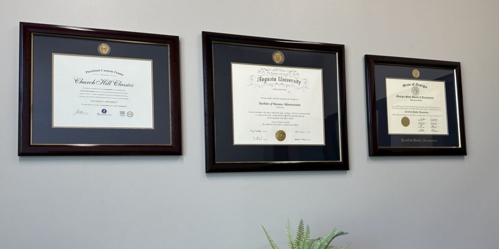 Three certificate frames with gold medallions hanging on a gray wall above a fern.