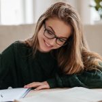Smiling woman with glasses sitting in living room working at table, writing in notebook and working on essay.
