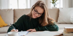 Smiling woman with glasses sitting in living room working at table, writing in notebook and working on essay.