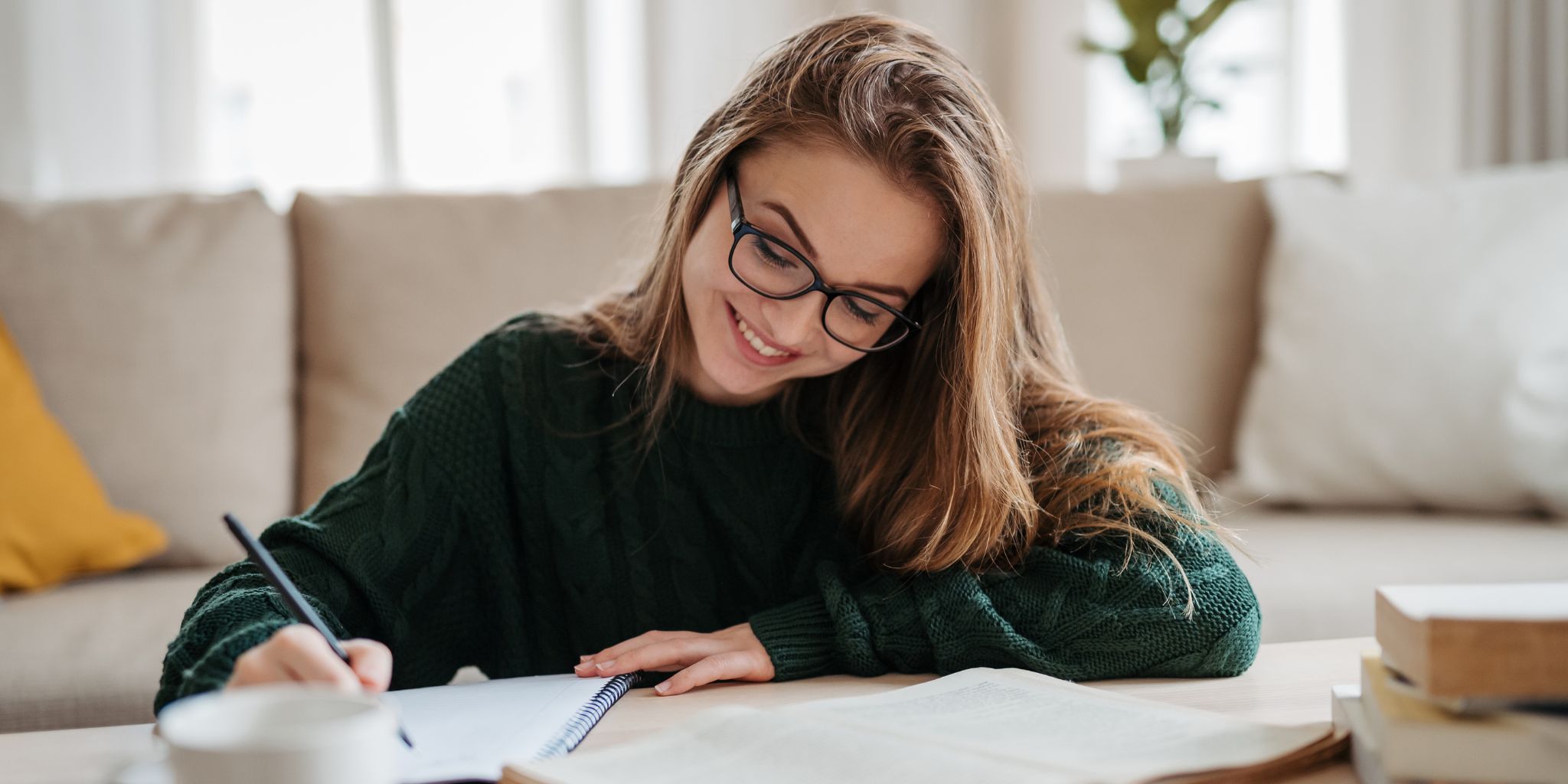 Smiling woman with glasses sitting in living room working at table, writing in notebook and working on essay.