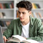 College student in library working on laptop with books in front of him.
