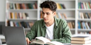 College student in library working on laptop with books in front of him.
