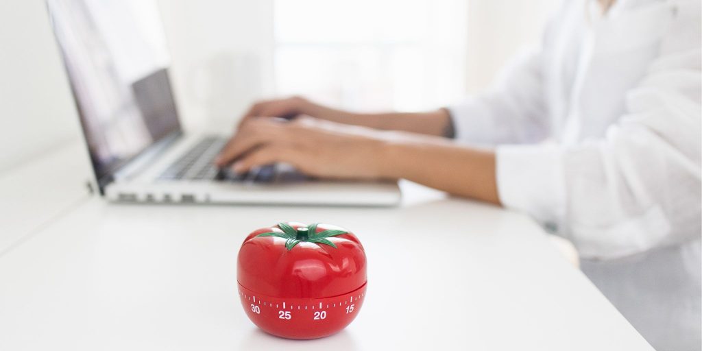 Closeup of a tomato-shaped timer on desk next to hands working on a laptop