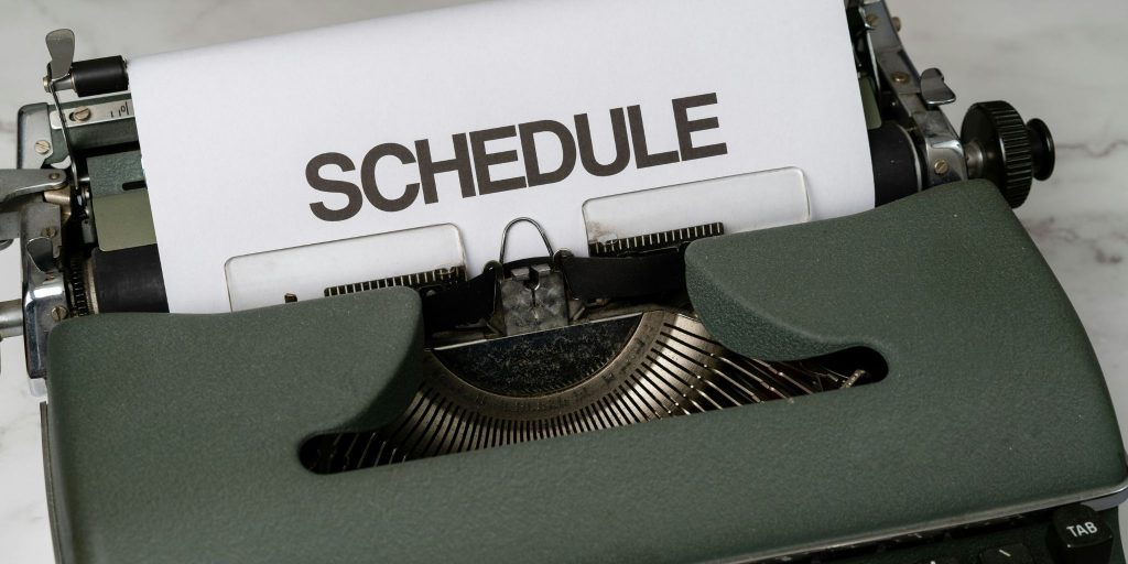Closeup of a dark green typewriter with a paper inserted in it that says SCHEDULE in all caps.