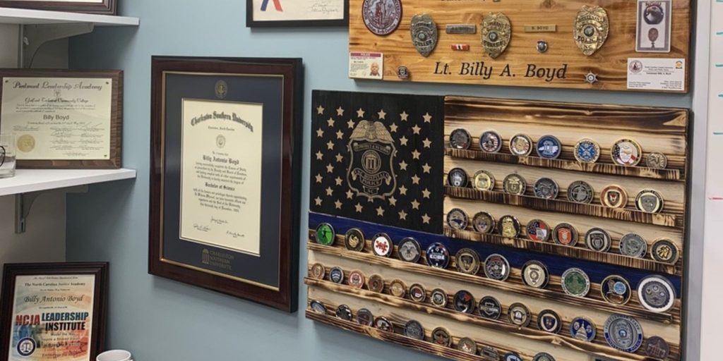 Office in police department with Charleston Southern University diploma frame next to wooden flag shaped challenge coin display and police awards on wall