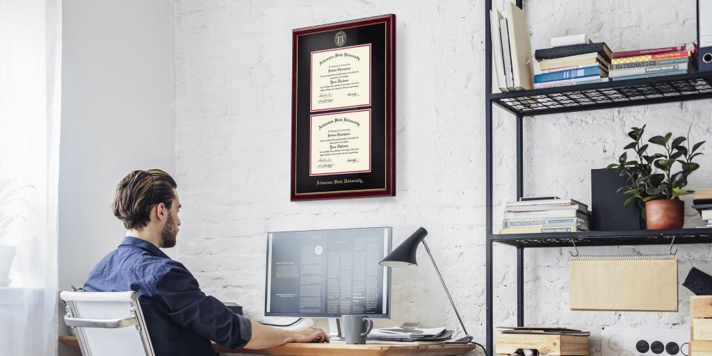 Man sitting at computer in office with an Arkansas State University double diploma frame on wall above his desk, which is next to a shelf.