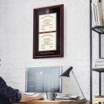 Man sitting at computer in office with an Arkansas State University double diploma frame on wall above his desk, which is next to a shelf.
