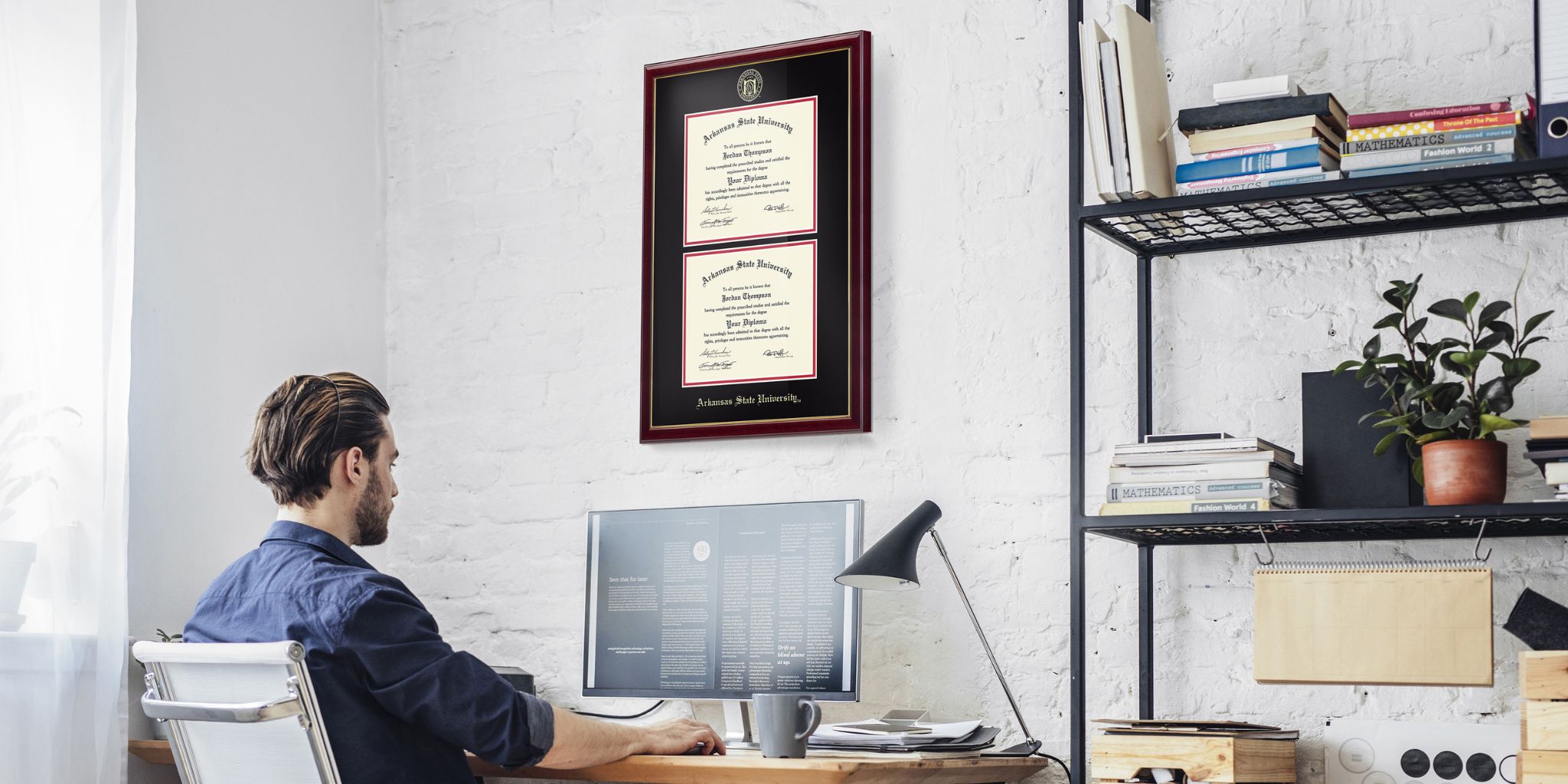 Man sitting at computer in office with an Arkansas State University double diploma frame on wall above his desk, which is next to a shelf.