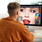 Person sitting at laptop in golden orange shirt working on colors on closeup of fashion model's face.