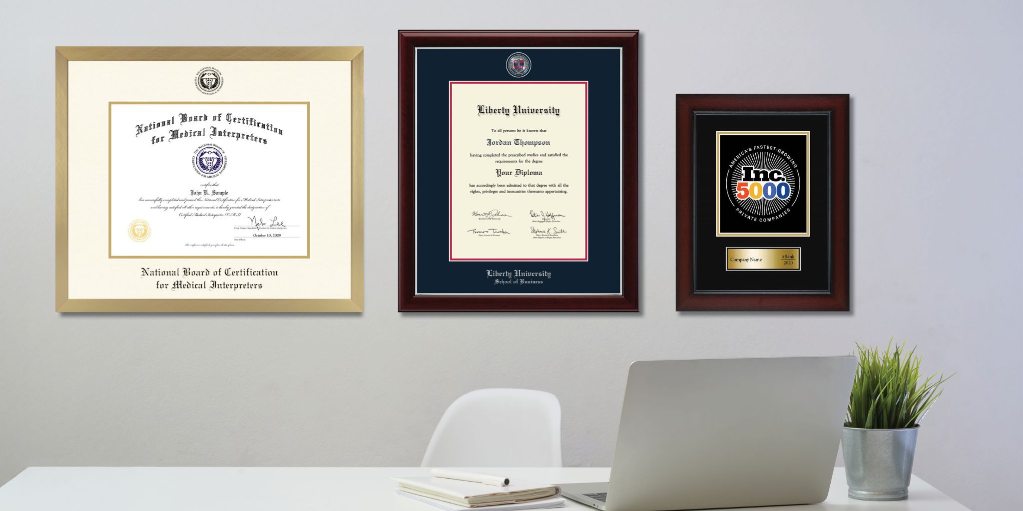 Gray office wall with medical certification frame, Liberty University dipoma frame, and Inc. 5000 award hanging above desk with chair, laptop and potted plant.