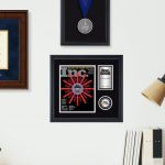 Office wall with a Penn State diploma frame, Inc. 5000 award, and medallion frame above a desk lamp, books, and a vase.