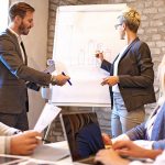 Two people leading meeting and holding markers as they write ideas on large paper pad.