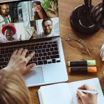 Overview of person working at desk with laptop showing four-way Zoom meeting. Headphones, notebook and plant on desk around laptop