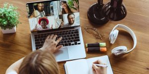 Overview of person working at desk with laptop showing four-way Zoom meeting. Headphones, notebook and plant on desk around laptop
