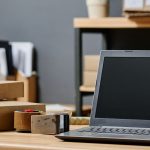 Laptop sitting atop a desk next to roll of shipping tape and cardboard shipping boxes.