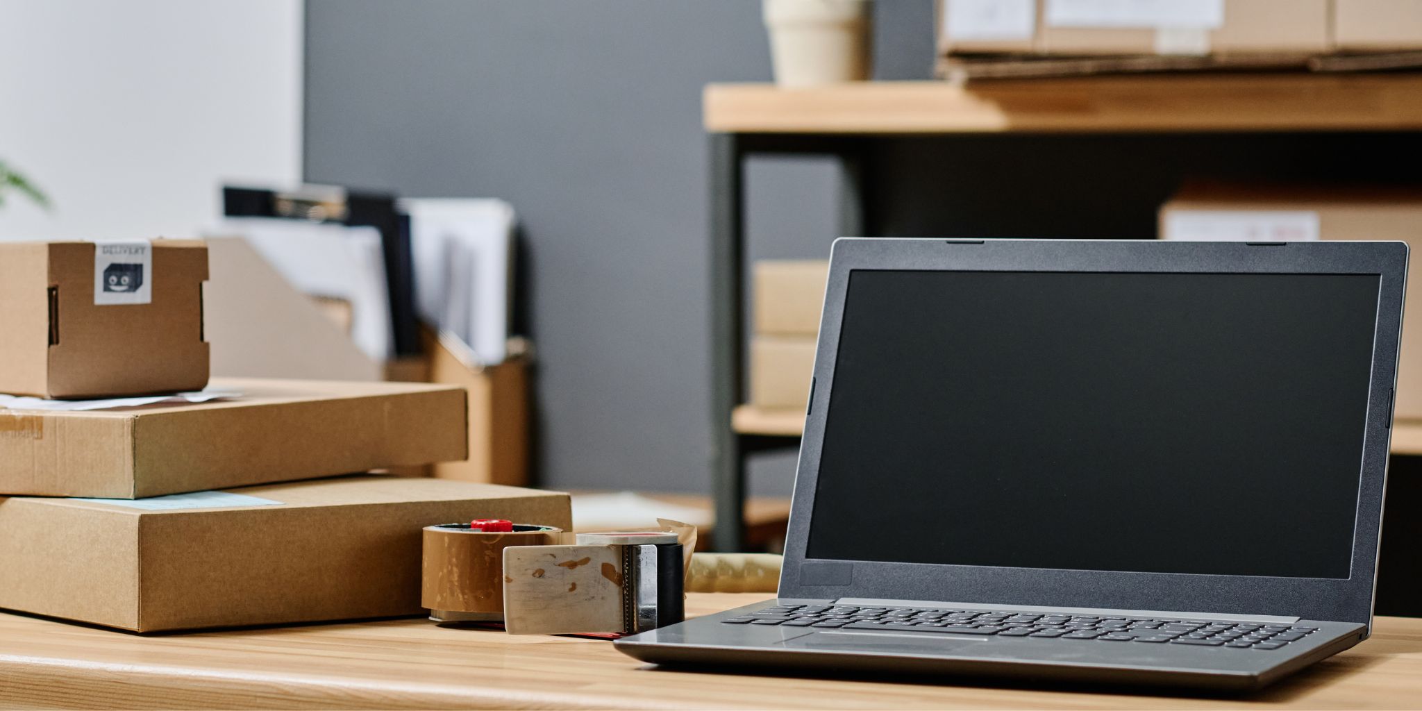 Laptop sitting atop a desk next to roll of shipping tape and cardboard shipping boxes.