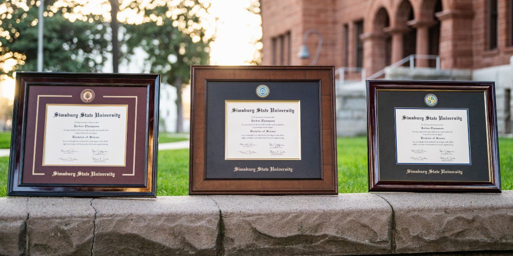 Three different styles of Simsbury State University diploma frames sitting atop a gray stone wall on a college campus.