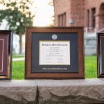Three different styles of Simsbury State University diploma frames sitting atop a gray stone wall on a college campus.
