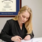 Blonde woman in suit in gray office working on papers on her desk with a University of Kentucky diploma frame on wall behind her.