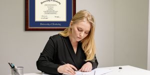 Blonde woman in suit in gray office working on papers on her desk with a University of Kentucky diploma frame on wall behind her.