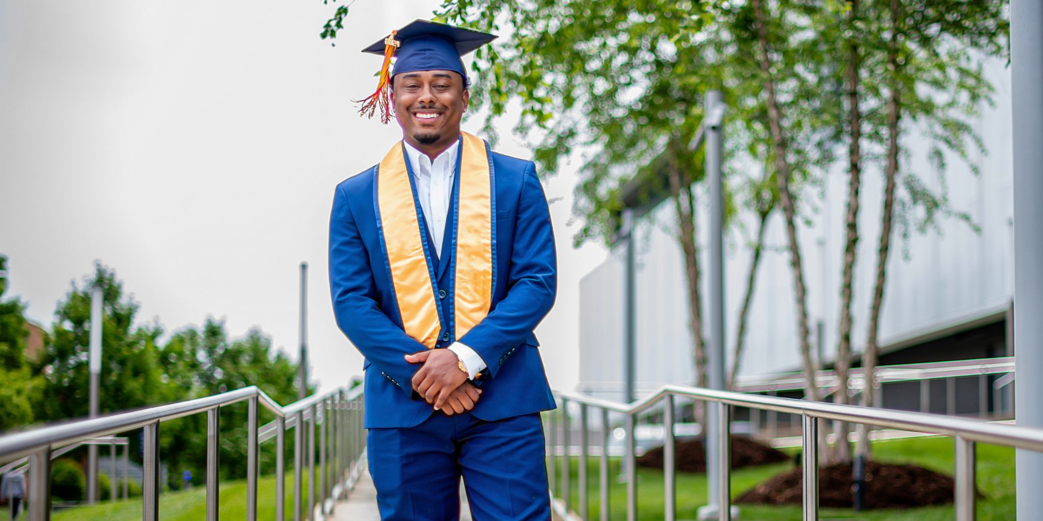 Smiling man in blue suit and blue graduation cap and yellow sash outdoors post-graduation.