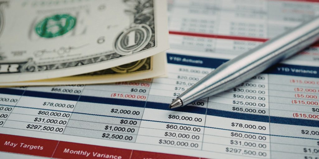 Closeup of budget spreadsheet with silver pen and $1 bills on top of it.