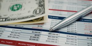 Closeup of budget spreadsheet with silver pen and $1 bills on top of it.