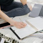 Closeup of person writing in notebook next to laptop on picnic blanket outdoors.
