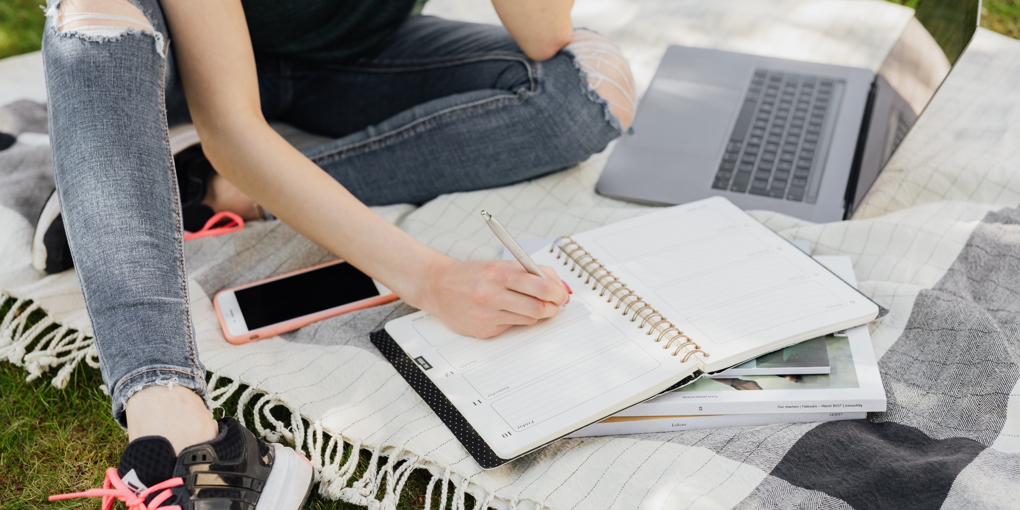 Closeup of person writing in notebook next to laptop on picnic blanket outdoors.