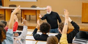 Professor with white beard in front of college lecture hall talking to students with their hands raised.