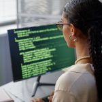 Back of person with long braids working on computer with two different screens filled with software coding.