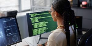 Back of person with long braids working on computer with two different screens filled with software coding.