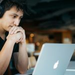 Man with headphones leaning his chin on clasped hands in cafe working on lap