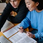 Two college students working side by side in front of laptop at desk with text books in front of them.