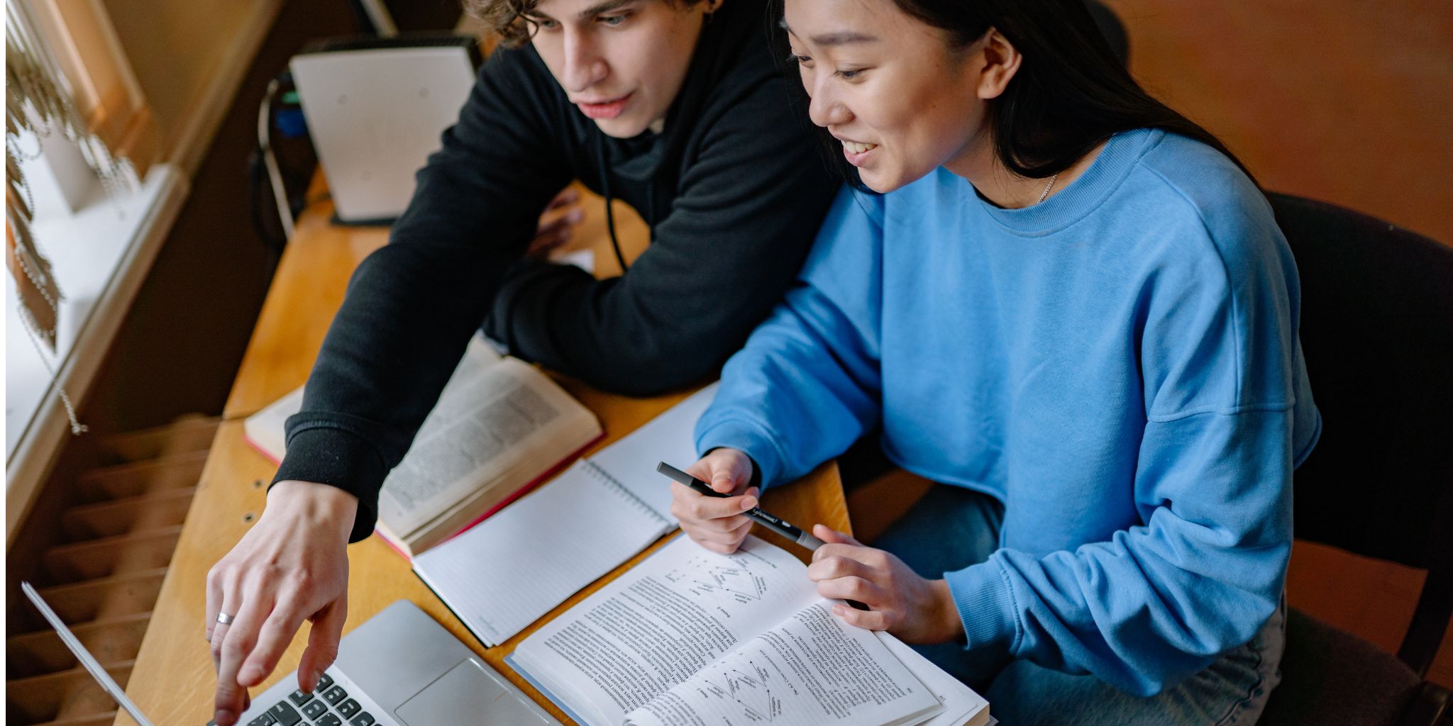 Two college students working side by side in front of laptop at desk with text books in front of them.