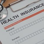 Closeup of a Health Insurance form on a clipboard with a stethoscope, pills, and pen nearby