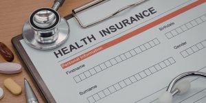 Closeup of a Health Insurance form on a clipboard with a stethoscope, pills, and pen nearby
