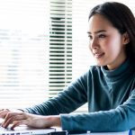 Woman in blue shirt working on laptop at desk in front of window with blinds.