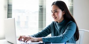 Woman in blue shirt working on laptop at desk in front of window with blinds.