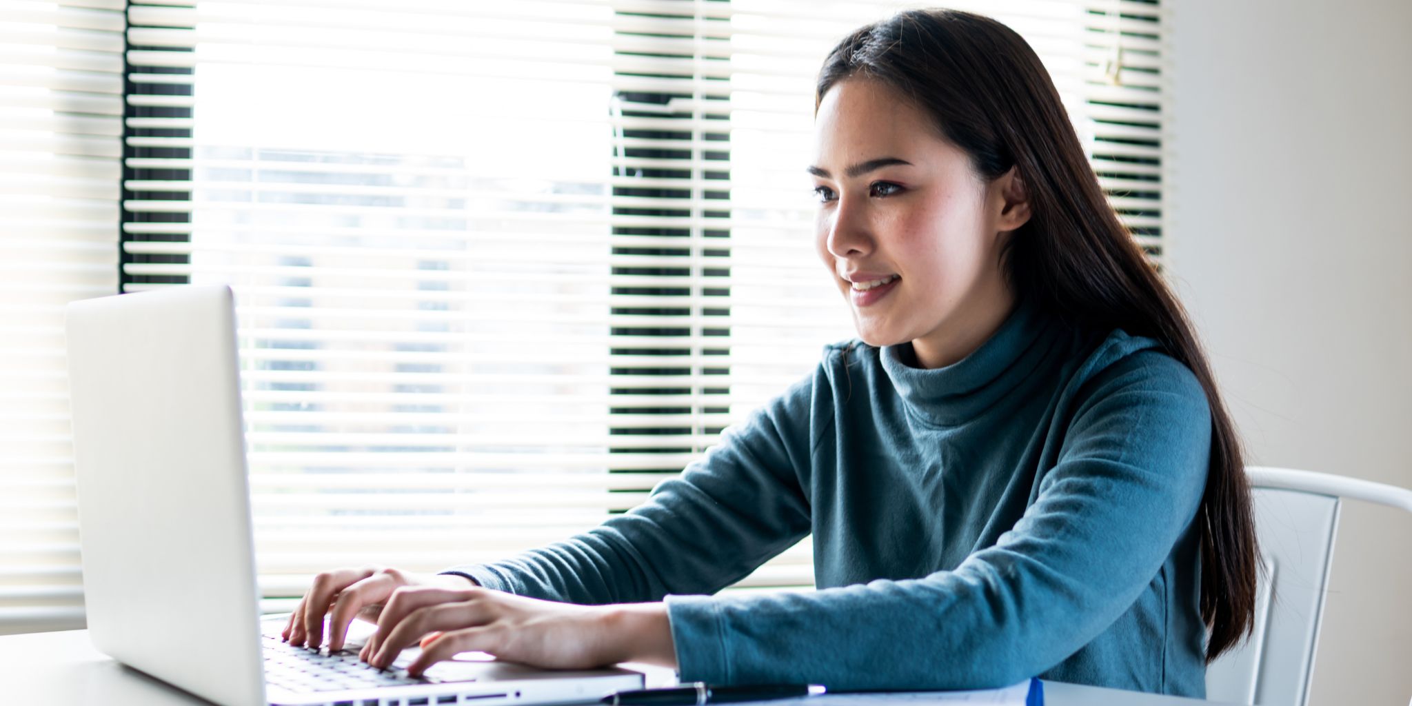 Woman in blue shirt working on laptop at desk in front of window with blinds.