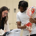Students looking at electronic medical device while one student holds anatomically correct skeleton.