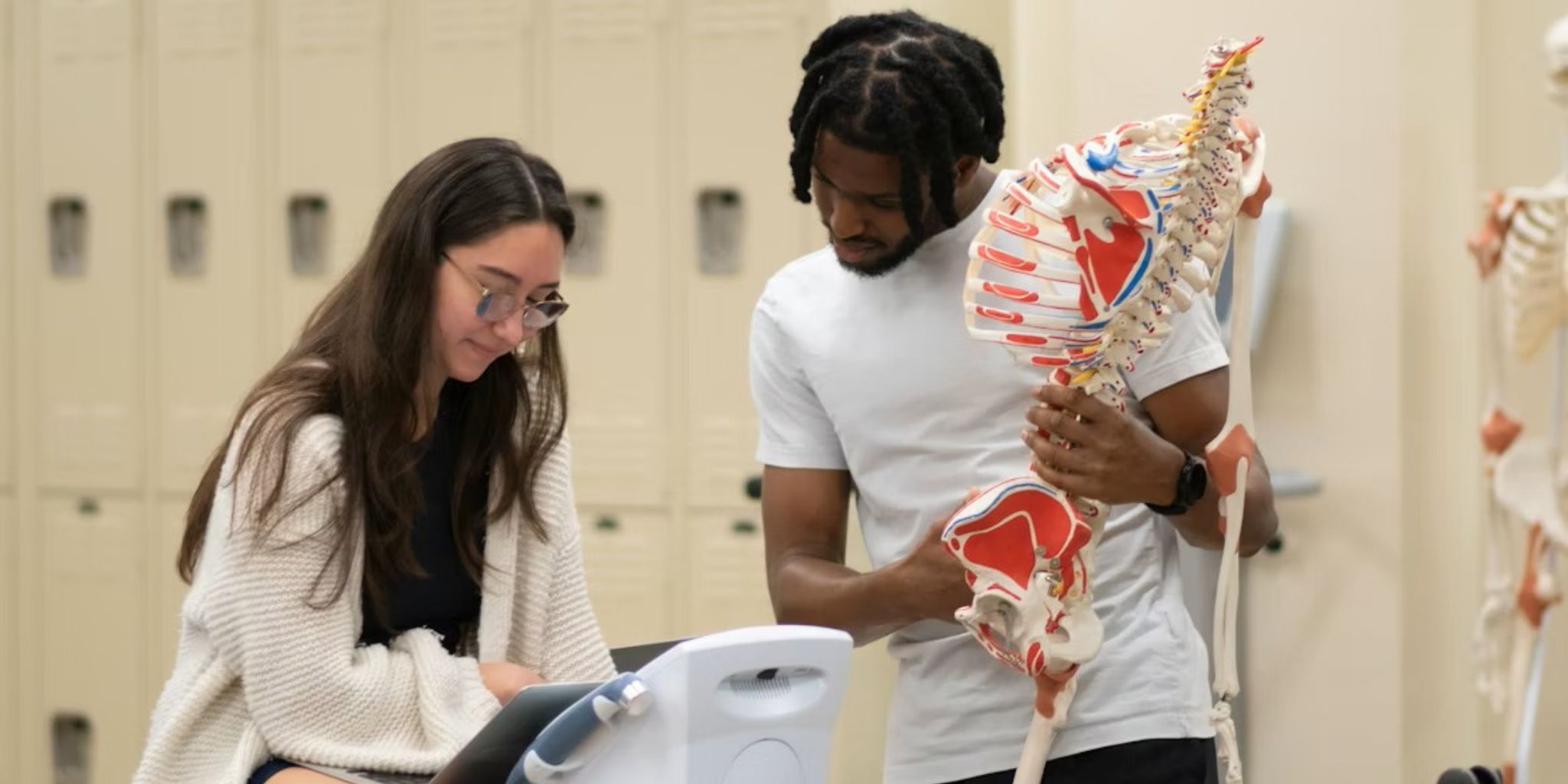 Students looking at electronic medical device while one student holds anatomically correct skeleton.